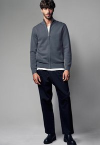 Grå stickad zip-up-jacka med strukturerat mönster, vit undertröja, svarta byxor och svarta stövlar. Modellen står mot en neutral bakgrund.