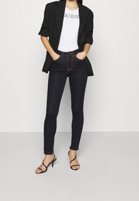 Jeans ajustados de cintura alta en denim oscuro, con costuras en contraste y cremallera, combinados con una blusa ajustada blanca y una chaqueta negra.