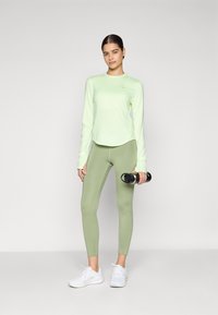Lys grøn langærmet sportstrøje med rund kant, parret med mid-rise grøn leggings. Hun holder en sort håndvægt og har hvide sneakers på.