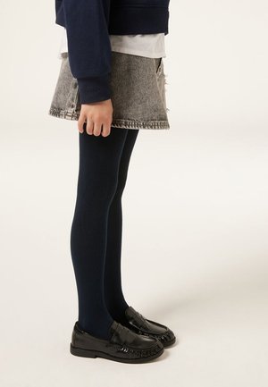 Kind trägt marineblaue Strumpfhosen, grauen Jeansrock, marineblauen Pullover und schwarze Lackleder-Slipper, steht vor einem schlichten hellen Hintergrund.