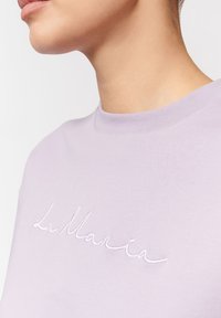 Zbliżenie na szyję i brodę osoby w jasnofioletowej koszuli z białym haftowanym tekstem na piersi.