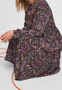 Blommig klänning i svart med ett flerfärgat mönster av små blommor. Långa ärmar, hög krage, flödande tyg och samlad detalj.