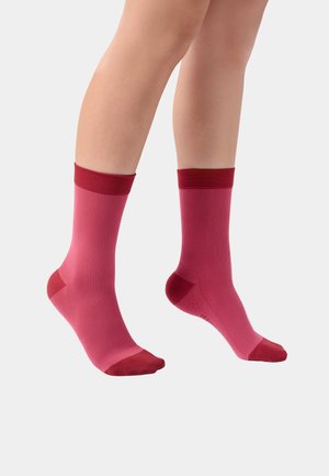 Beine mit gerippten Kniestrümpfen in Dunkelrot an Bündchen, Zehen und Fersen vor einem schlichten, hellen Hintergrund.