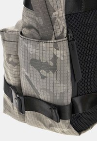 Rucksack aus grauem Camouflage-Stoff mit strukturierter Oberfläche. Enthält einen schwarzen Mesh-Bereich, Nylonträger und eine Seitentasche mit Clip.