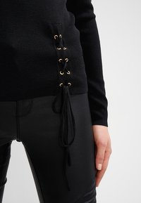 Zwarte gebreide top met zijkant vetersluiting, voorzien van gouden oogjes en losse strikjes, gecombineerd met zwarte neplederen broek.