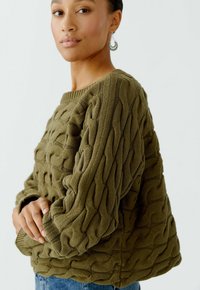 Olijfgroen kabelgebreid sweater met een textuurpatroon, ronde hals en een losse pasvorm, gecombineerd met blauwe spijkerbroek.
