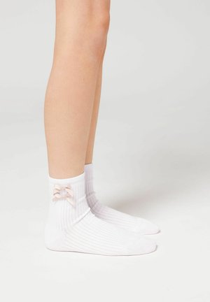 Calze bianche a coste con un dettaglio di fiocco rosa sulla parte esterna della caviglia. Il tessuto sembra morbido ed elastico, progettato per una lunghezza fino alla caviglia.