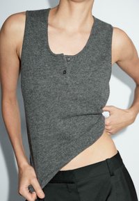 Top senza maniche grigio in maglia con scollatura a barca e dettagli con bottoni. Texture morbida e vestibilità rilassata, abbinato a pantaloni neri a vita alta.