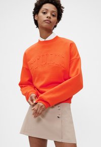 Oranžová mikina s texturovaným nápisem "STUDIO", kulatým výstřihem a dlouhými rukávy, doplněná béžovou mini sukní s knoflíkovými detaily.