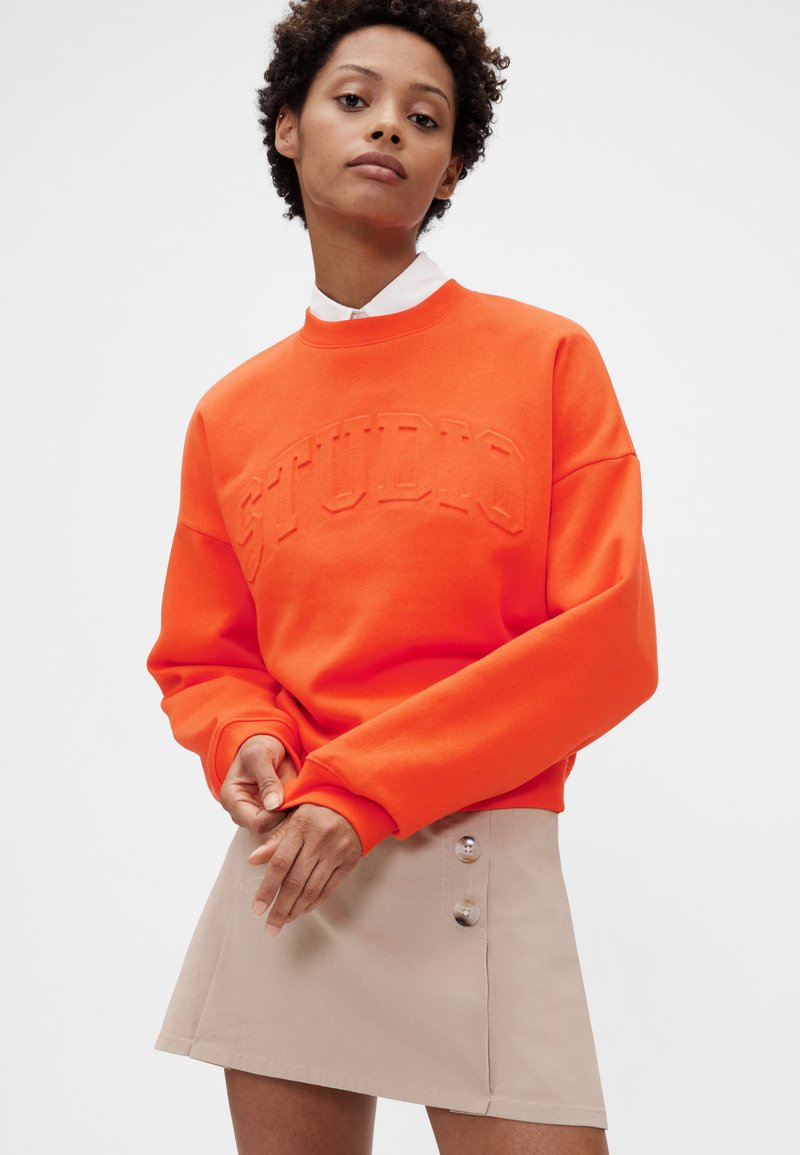 Oranžová mikina s texturovaným nápisem "STUDIO", kulatým výstřihem a dlouhými rukávy, doplněná béžovou mini sukní s knoflíkovými detaily.