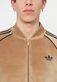 Hnědá sametová bunda na zip s projektem na límci, vyznačující se černými pruhy a černým trojlístkem na levé straně. Hladká textura.