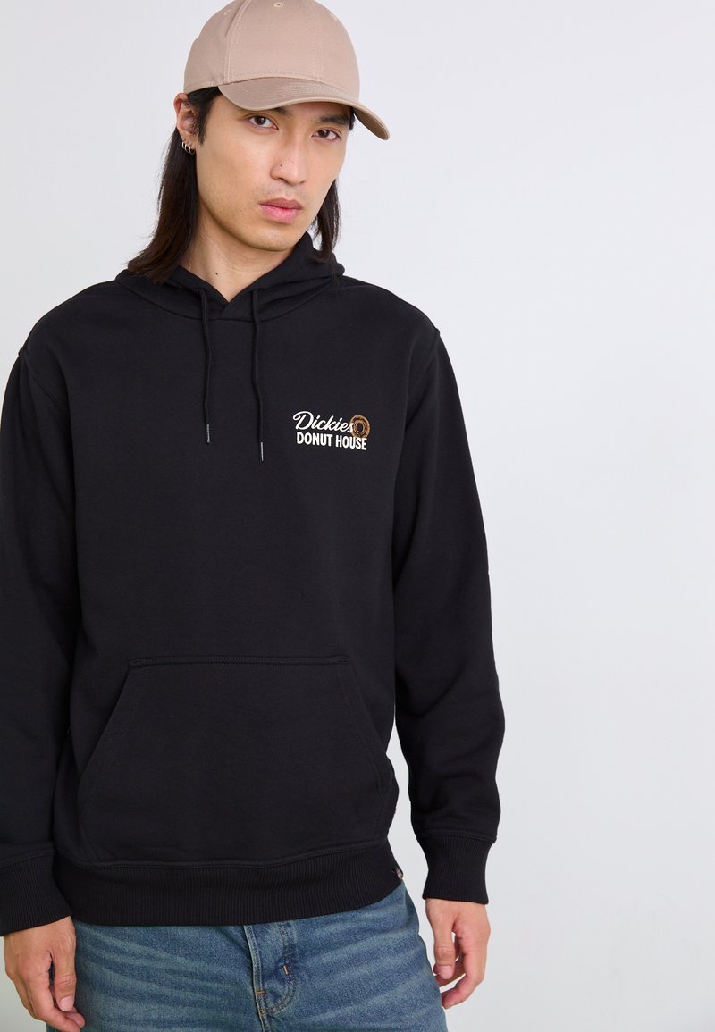 Felpa nera con tasca a marsupio, con un logo "Dickies Donut House" bianco e marrone. Abbinata a un cappellino beige chiaro.