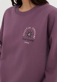 Paarse katoenen sweatshirt met een ronde halslijn en een vooropdruk met de tekst "Celestial Symphony" en "Solar Eclipse," aangevuld met ontwerpelementen.