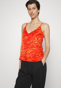 Top vermelho sem mangas com decote em V, apresentando um padrão de listras de zebra texturizadas em tons de laranja. Tecido leve, combinado com calças pretas.