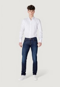 Camisa blanca de manga larga con botones, jeans de mezclilla en azul oscuro y zapatillas blancas. Corte clásico, tela suave y detalles de costura visibles.
