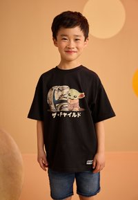 Camiseta negra de algodón con estampado gráfico de un personaje y un casco; combinada con pantalones cortos de mezclilla. Diseño sencillo con cuello redondo.