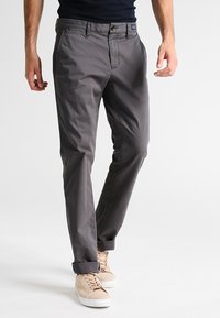 Mężczyzna w ciemnoszarych chinosach z mankietami i beżowych sneakersach, stojący na tle jednolitego jasnego tła.