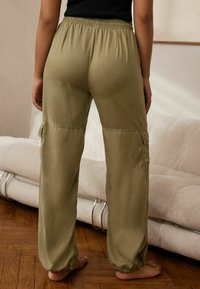 Persoon in losse olijfgroene cargobroek met elastische tailleband en boorden, staande op blote voeten op een houten vloer nabij een beige bank.