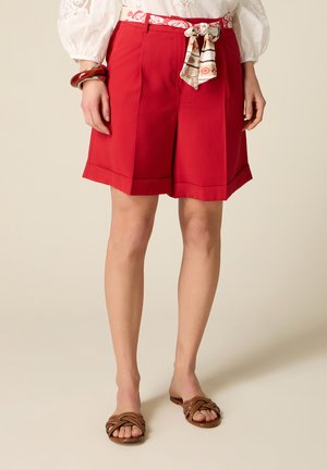 Kobieta nosząca czerwone szorty do kolan z podwiniętymi brzegami, wzorzysty materiałowy pasek, białą bluzkę, brązowe plecione sandały i masywną bransoletkę.