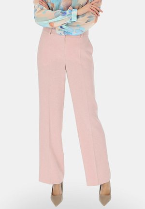 Kobieta w przezroczystej, kwiatowej bluzce wpuszczonej w spodnie o wysokim stanie, szerokich nogawkach w jasnoróżowym kolorze oraz beżowych butach na szpic, ze skrzyżowanymi ramionami.