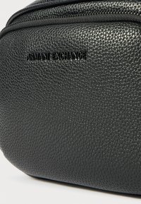Crna torba od teksturirane kože s utisnutim logotipom "ARMANI EXCHANGE" koji se ističe sprijeda. Glatko zatvaranje na zatvarač.