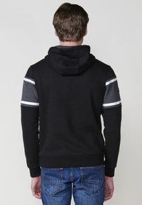 Model masculin văzut din spate, purtând un hanorac negru cu glugă, cu mânecile în dungi gri și alb, asortat cu blugi blue.
