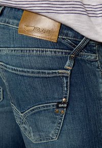 Jeans de mezclilla en azul oscuro con un acabado desgastado. Presenta una etiqueta de cuero marrón con "GAS" y una pequeña etiqueta negra en el bolsillo. Remaches metálicos presentes.