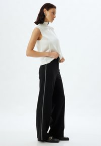 Witte mouwloze top met hoge hals, gecombineerd met zwarte wijde broek met witte zijstrepen en platte zwarte schoenen.