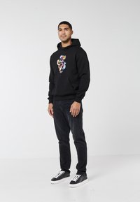 Svart hoodie med en färgstark grafisk design på bröstet, kombinerad med mörka jeans och svarta sneakers med vita snören och sula.