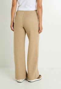 Pantalones acampanados beige en una tela texturizada. Cuentan con una cintura alta y piernas anchas. Combinados con una blusa blanca y zapatillas marrones.