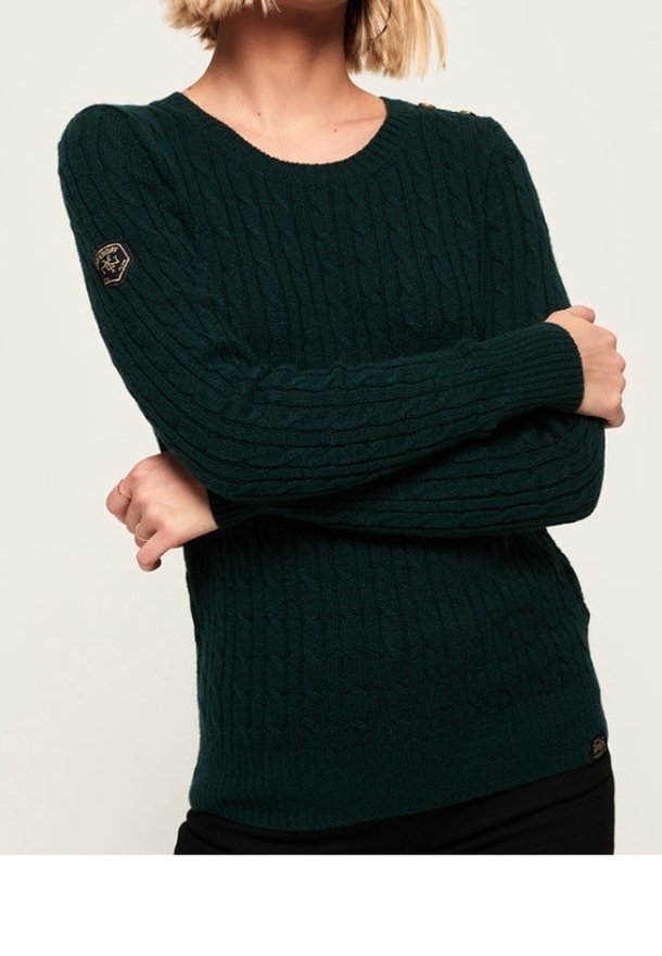 Grüner strukturierter Pullover mit Rundhalsausschnitt, der ein Zopfmuster und ein gesticktes Logo am linken Ärmel aufweist.