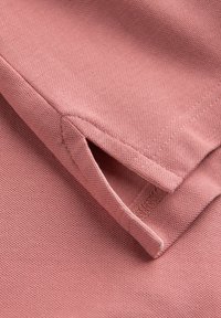 Rosa bomullstyg i närbild med en strukturerad väv. Skjortan har en rundad nederkant med sidoflikar och rena, sydda kanter.