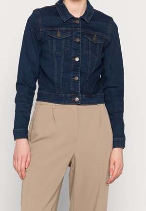 Kvinde iført en mørkeblå denimjakke med knaplukning og beige plisserede højttaljede bukser, stående mod en hvid baggrund.