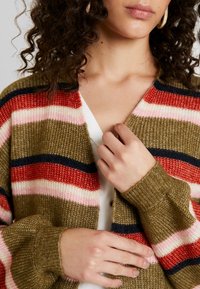 Kaki cardigan s texturovaným vzorem, který obsahuje vodorovné pruhy v červené, růžové a námořnické modři. Výstřih do V a žebrované manžety. Zapínání na knoflíky na přední straně.