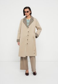 Trench-coat beige avec un col et des poignets pied-de-poule, doté d'une fermeture à boutons sur le devant et d'une ceinture nouée à la taille. Porté avec un pantalon large et clair.