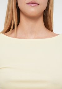 Ανοιχτό κίτρινο ριμπ πλεκτό τοπ με φαρδύ λαιμό, που δείχνει ομαλή υφή και εφαρμοστή σχεδίαση, τονίζοντας το κόκαλο του λαιμού και τους ώμους.