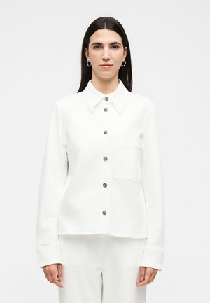 Woman with long dark hair wearing a white buttoned shirt with a chest pocket and matching white pants, standing against a plain background.