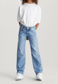 Camicia bianca a maniche corte e jeans ampi azzurri. I jeans presentano un bottone e tasche, mentre le scarpe sono sneakers bianche.