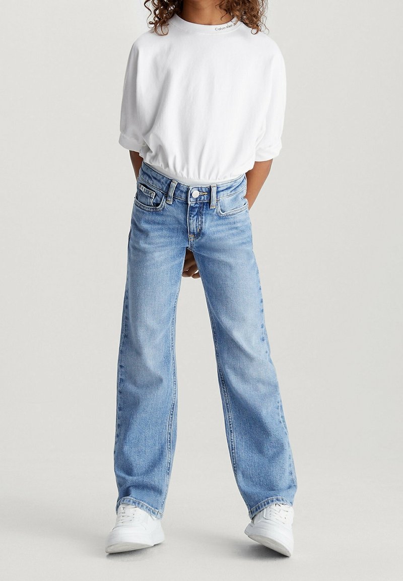 Camicia bianca a maniche corte e jeans ampi azzurri. I jeans presentano un bottone e tasche, mentre le scarpe sono sneakers bianche.