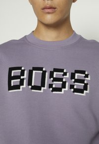 Gros plan sur une personne portant un sweat-shirt violet clair avec des lettres pixellisées noires et blanches formant le mot « BOSS » sur la poitrine.