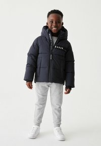 Doudoune navy à capuche, avec une fermeture éclair et un logo sur la poitrine. Associée à un pantalon de survêtement gris clair et des baskets blanches.
