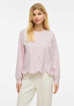 Lichtroze cropped sweatshirt met gegolfde zoom en textuurdetails. Lange mouwen, ronde hals, zachte stof, gecombineerd met crèmekleurige broeken.