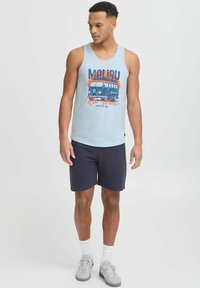 Világoskék ujjatlan felső, rajta "Malibu" felirat és egy vintage kisbusz design. Sötét rövidnadrággal és fehér zoknikkal párosítva szürke sportcipőt visel.