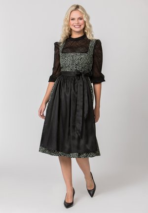 Junge Frau lächelt, trägt eine schwarze transparente Bluse und ein grünes Kleid mit Leopardenmuster und eine schwarze Satin-Schürze, die an der Taille gebunden ist.