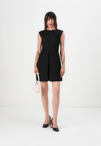 Black quilted dress with cap sleeves, pleated front, and two pockets; features a fabric flower accent. Paired with a small pink handbag.
