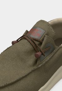 Uformell olivengrønn slip-on-sko med teksturert stoff, elastiske lisser med røde prikker og lysfarget såle.