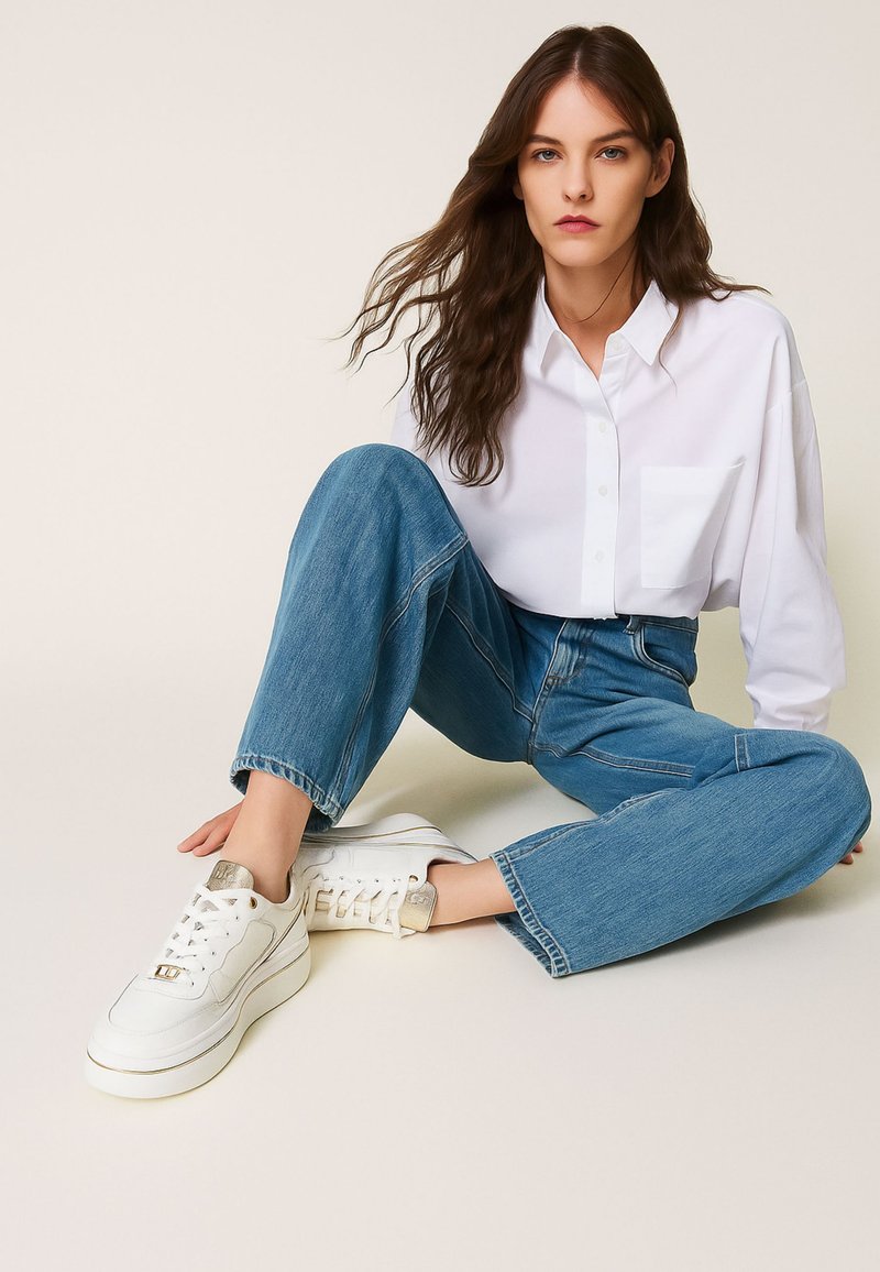 Chemise blanche à boutons, jeans bleus taille haute et baskets blanches avec accents dorés. Look décontracté avec une silhouette relaxée.