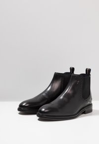 Las botas Chelsea de cuero negro presentan un acabado suave, paneles laterales elásticos y un detalle decorativo de perforaciones en la parte superior. Suelas planas.