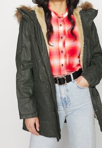 Olivgrön parkas med fuskpälsfodrad huva, knappstängning, flera fickor, lagd över en röd rutig skjorta, tillsammans med högmidjade jeans.
