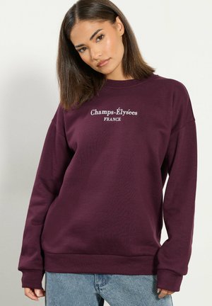 Młoda kobieta z prostymi brązowymi włosami nosi fioletową bluzę z napisem „Champs-Élysées FRANCE” oraz jasnoniebieskie jeansy, stoi na białym tle.
